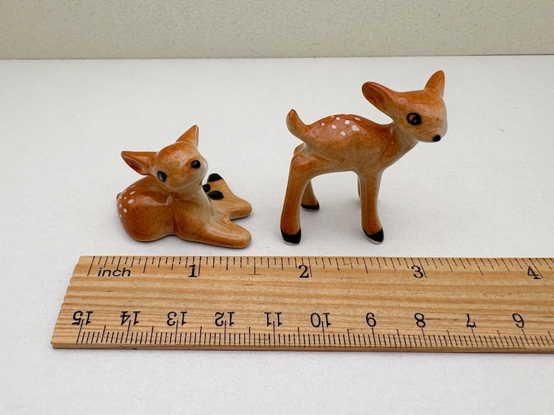 May include: Two ceramic deer figurines. One is lying down, the other is standing. Both are brown with white spots and black accents. A wooden ruler with inch markings is in the foreground.