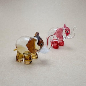 May include: Two glass elephant ornaments. One is amber and brown, the other is red and clear. Both have detailed trunks, ears, and legs. The elephants are small decorative items, suitable for display.
