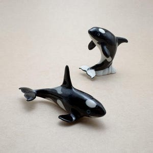 May include: Two ceramic orca figurines. One orca is lying down, and the other is leaping. Both have black and white markings, with a dorsal fin and tail. The figurines are on a neutral background.
