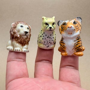 Puede incluir: Tres marionetas de dedo de cerámica que representan un león con melena marrón, un guepardo amarillo con manchas negras y un tigre naranja con rayas negras. Cada figura tiene rasgos pintados y una base hueca para colocar el dedo.