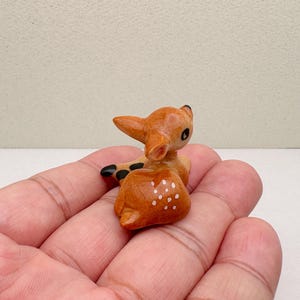 May include: A small, wooden figurine of a fawn, lying down with its head turned. The fawn is light brown with white spots on its back and black accents on its ears and hooves. The figurine is held in a person's hand.