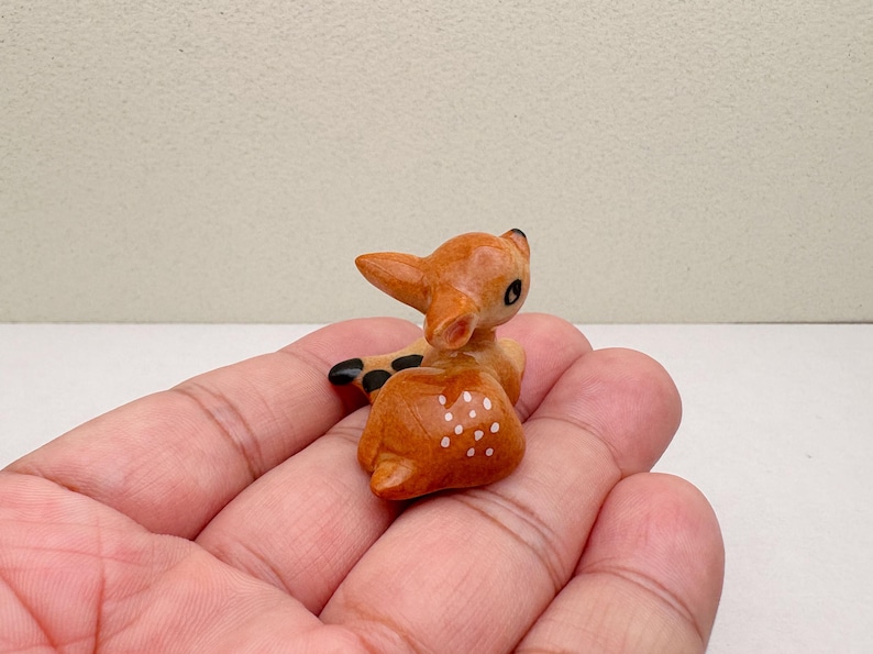 May include: A small, wooden figurine of a fawn, lying down with its head turned. The fawn is light brown with white spots on its back and black accents on its ears and hooves. The figurine is held in a person's hand.