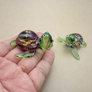 Puede incluir: Dos figuras de tortugas de cristal en miniatura. Una tortuga tiene un caparazón morado y dorado con aletas y cabeza verdes. La otra tortuga tiene un caparazón verde con una mezcla de colores y aletas verdes. Ambas tienen ojos blancos.