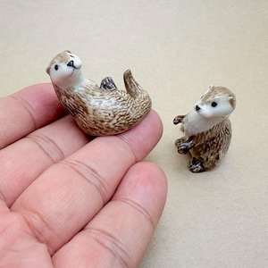 Könnte beinhalten: Zwei kleine Otterfiguren aus Keramik mit glänzendem braun-weißem Fellmuster. Ein Otter liegt auf dem Rücken mit eingerolltem Schwanz, während der andere Otter auf seinen Hinterbeinen steht und die Vorderpfoten nach vorne gestreckt hält.