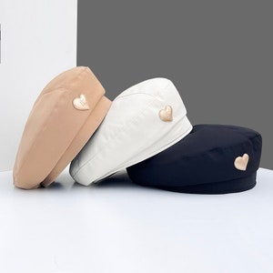 Op de afbeelding: Drie baretten in verschillende kleuren: beige, wit en zwart. Elke baret heeft een gouden hartvormig embleem.