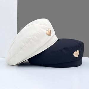 Op de afbeelding: Twee baretten, een witte en een zwarte, met gouden hart accenten. De witte baret heeft een gouden hart aan de zijkant, en de zwarte baret heeft een gouden hart aan de achterkant.