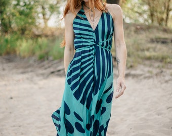 May include: A woman wearing a blue and black patterned maxi dress with a halter neckline. The dress has a geometric design with stripes and circles. She is standing on a sandy beach.