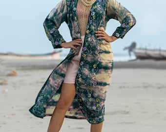 May include: A woman wearing a long, green and white floral patterned kimono with a pink skirt and gold sandals. She is standing on a sandy beach.