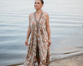 May include: A woman wearing a floral maxi dress stands on a sandy beach with a body of water in the background.