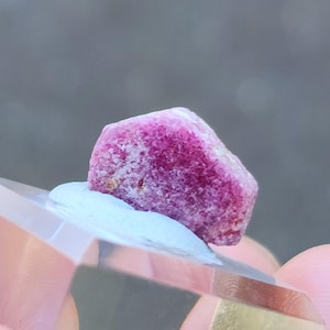 Puede incluir: Un cristal de rubí rosa intenso con una superficie rugosa y natural. El cristal está colocado sobre un soporte de exhibición de acrílico transparente.