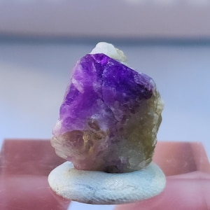May include: A purple and green crystal with a rough, natural texture. The crystal is sitting on a white surface.