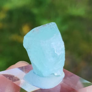May include: A light blue aquamarine crystal with a rough, natural texture. The crystal is sitting on a white base.