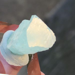 May include: A light blue crystal with a translucent, almost white, centre. The crystal is sitting on a light blue surface.