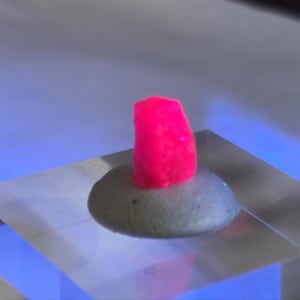 May include: A vibrant pink, rectangular-shaped object sits atop a gray, rounded base. The base has a textured surface. The objects are displayed on a clear, square platform, with a blue and white background.