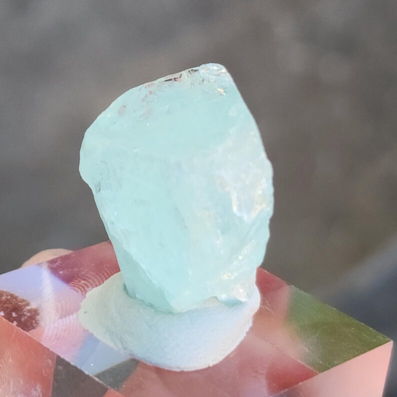May include: A light blue aquamarine crystal with a rough, natural shape. The crystal is sitting on a white display stand.