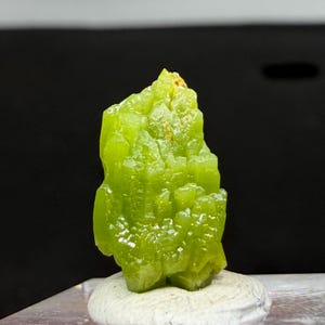Puede incluir: Un vibrante espécimen mineral de color verde lima con una estructura cristalina. El mineral es de un verde brillante y translúcido, con una textura ligeramente rugosa. Se asienta sobre una pequeña base de color claro, con un fondo oscuro.