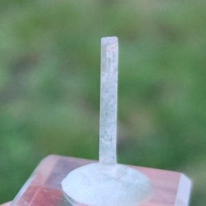 May include: A clear, rectangular crystal stands upright on a light blue, rounded base. The crystal is translucent with a slightly textured surface. The background is a blurred green, suggesting an outdoor setting.