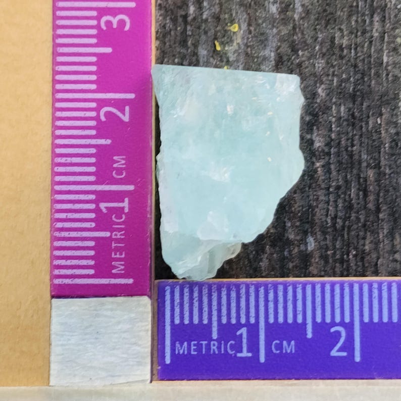 May include: A light blue aquamarine crystal with a rough, natural texture. The crystal is approximately 1 cm in size and is measured against a ruler.