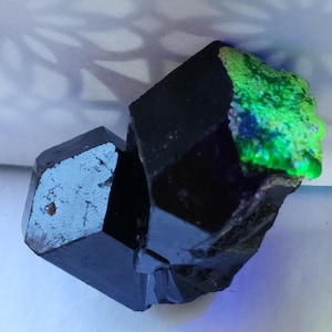 May include: A black crystal with a green and blue iridescent sheen. The crystal is faceted and has a sharp, geometric shape.