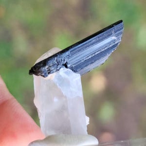 May include: A black tourmaline crystal point atop a clear quartz crystal. The tourmaline is striated and has a sharp point. The quartz is clear and has a smooth surface.