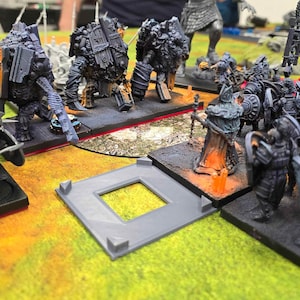 May include: A tabletop wargaming scene with a variety of miniature figures, including a group of armored warriors, a large mechanical creature, and a smaller figure with a staff. The figures are arranged on a green and brown gaming mat with a gray square in the center.