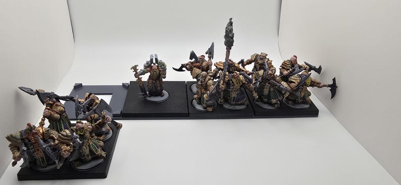May include: A set of 12 painted miniature figures, depicting warriors with axes and shields, standing on black bases. The figures are in various poses, some holding weapons, others with shields raised. The figures are painted in shades of brown, green, and black.
