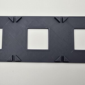 May include: A dark gray plastic frame with three rectangular openings. The frame has a textured surface and is designed to hold three items.