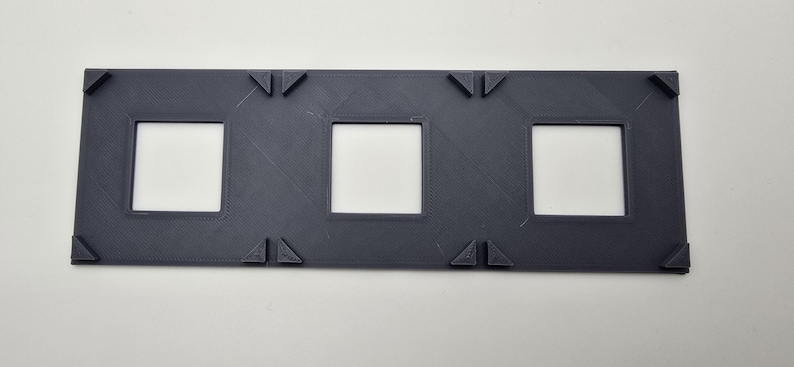 May include: A dark gray plastic frame with three rectangular openings. The frame has a textured surface and is designed to hold three items.