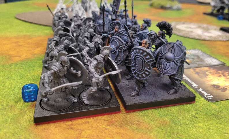 May include: A set of two miniature plastic army figures, one in silver and one in dark gray, with shields and swords. The figures are on black bases with red trim. The figures are arranged in a battle formation.