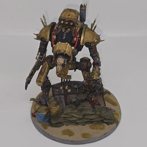 May include: A gold and brown painted miniature robot figure with a skull-like head, standing on a circular base with sand and green painted rocks. The robot is holding a weapon in its right hand and has a large, detailed weapon on its back.