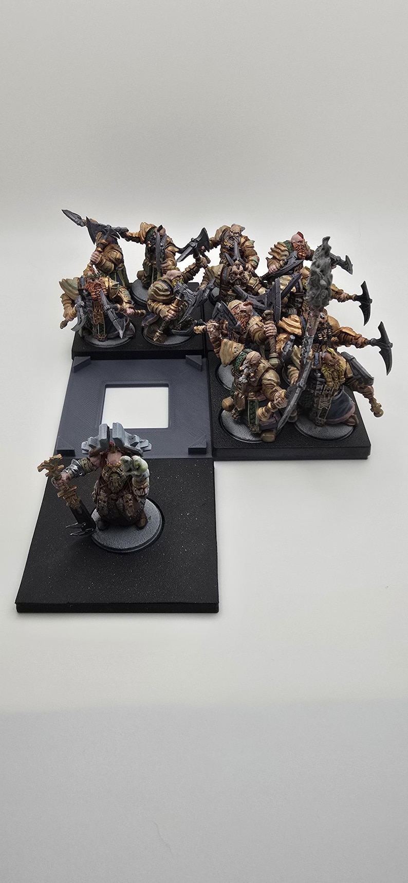 May include: A group of 10 painted miniature figures of dwarves, each holding a weapon and standing on a black base. The figures are arranged in a semi-circle formation, with one figure standing in front of the others. The figures are all wearing armor and have beards.
