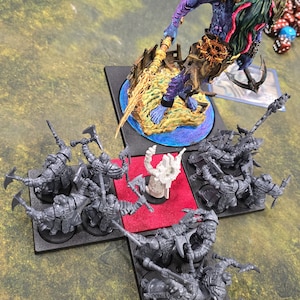 May include: A tabletop game scene featuring miniature figures. A large, purple, tentacled creature with a golden weapon stands above several gray figures with axes. The figures are arranged on black bases, with a red and blue playing surface.