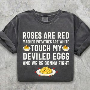 May include: A charcoal gray short-sleeve t-shirt featuring the white text: ROSES ARE RED MASHED POTATOES ARE WHITE TOUCH MY DEVILED EGGS AND WE'RE GONNA FIGHT. The design includes illustrations of deviled eggs on plates with small red hearts.