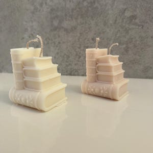 May include: Two ivory-colored candles shaped like stacks of books. Each candle has a wick and is detailed with embossed floral and spine designs. The candles are on a white surface against a gray background.