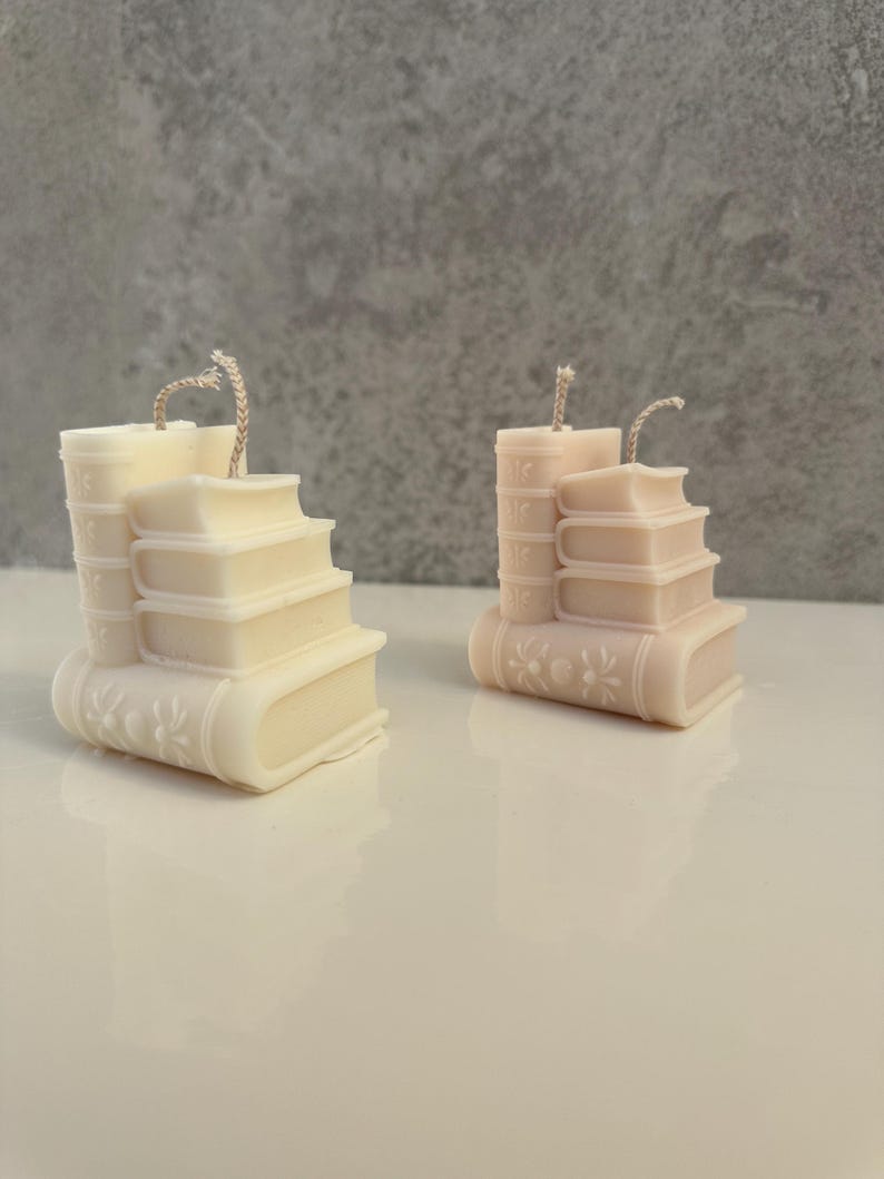 May include: Two ivory-colored candles shaped like stacks of books. Each candle has a wick and is detailed with embossed floral and spine designs. The candles are on a white surface against a gray background.