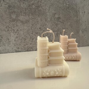 May include: Ivory-colored candle shaped like a stack of books, with a decorative floral design on the bottom book. The candle has a rustic twine wick. Another candle in a light pink hue is also visible.