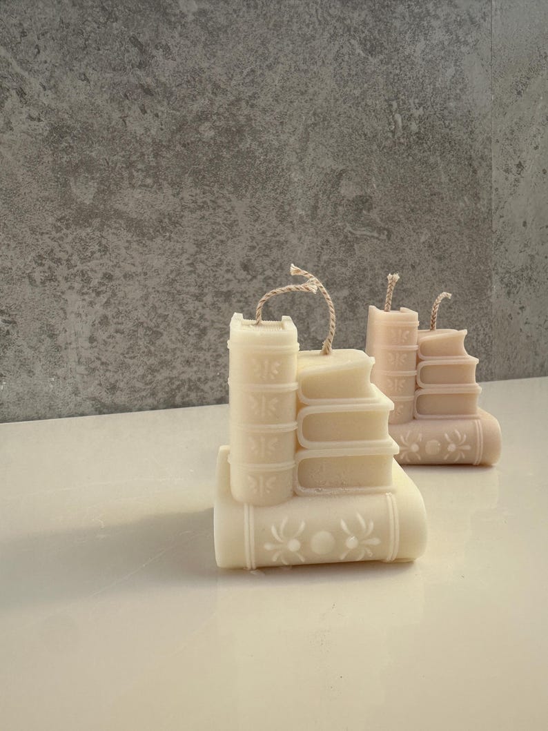 May include: Ivory-colored candle shaped like a stack of books, with a decorative floral design on the bottom book. The candle has a rustic twine wick. Another candle in a light pink hue is also visible.