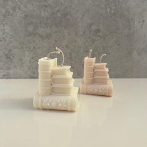 May include: Two decorative candles shaped like stacks of books. The candles are in shades of cream and peach, with detailed book spines and floral accents. Each candle has a wick extending from the top, suggesting they are ready to be lit.