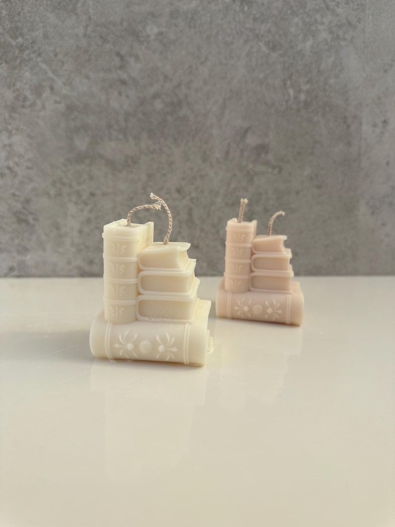May include: Two decorative candles shaped like stacks of books. The candles are in shades of cream and peach, with detailed book spines and floral accents. Each candle has a wick extending from the top, suggesting they are ready to be lit.
