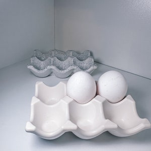 Könnte beinhalten: Zwei weiße Eier in einem weißen Keramik-Eierkarton. Der Karton hat sechs Fächer und steht auf einer weißen Oberfläche.