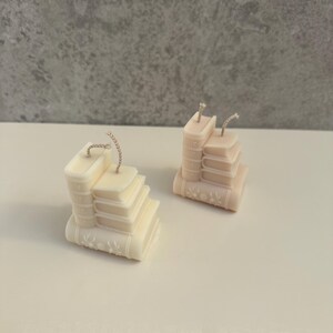May include: Two ivory-colored candles shaped like stacks of books. Each candle has a wick and is detailed with embossed book spines and floral designs. The candles are on a white surface against a gray background.