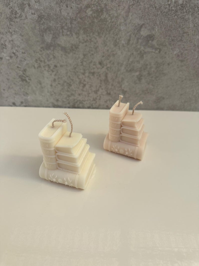 May include: Two ivory-colored candles shaped like stacks of books. Each candle has a wick and is detailed with embossed book spines and floral designs. The candles are on a white surface against a gray background.