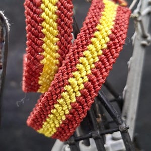May include: A woven friendship bracelet featuring a central yellow stripe flanked by two wider red stripes. The textile accessory is constructed from knotted thread, displaying a consistent diagonal pattern throughout the length of the band.