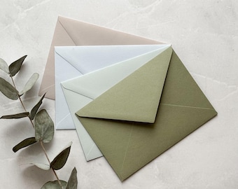 May include: Four square envelopes in beige, white, and olive green are layered on a light grey marble surface. A sprig of eucalyptus is in the corner. The envelopes are in a variety of muted tones.