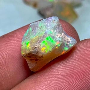 May include: A rough opal gemstone with vibrant colors of green, yellow, and blue. The gemstone is held in a hand, showcasing its natural, unpolished texture. Other opals are visible in the background.
