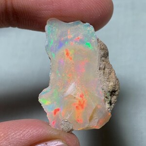 May include: A rough-cut opal gemstone with a vibrant display of colors. The opal features flashes of red, orange, green, and blue within a milky white base. The stone is held between fingers, showcasing its natural, irregular shape.