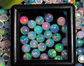 May include: A close-up of a small box filled with iridescent opal gemstones. The opals are round and have a variety of colours, including green, blue, red, and purple. The box is black and has a clear lid.