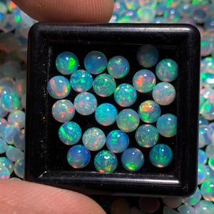 Op de afbeelding: Een close-up van een verzameling ronde, iriserende opaal edelstenen in een zwarte lade. De opalen zijn in verschillende tinten blauw, groen en wit, met kleurflitsen.