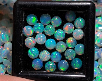 May include: A close-up of a collection of round, iridescent opal gemstones in a black tray. The opals are various shades of blue, green, and white, with flashes of colour.
