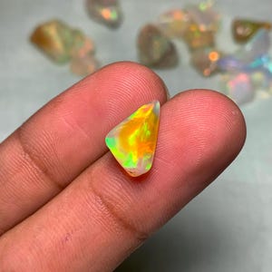 May include: A vibrant, triangular opal gemstone with a mix of colors, including orange, yellow, and green, is held between fingers. The opal has a polished surface and displays a play of light. Other opals are visible in the background.